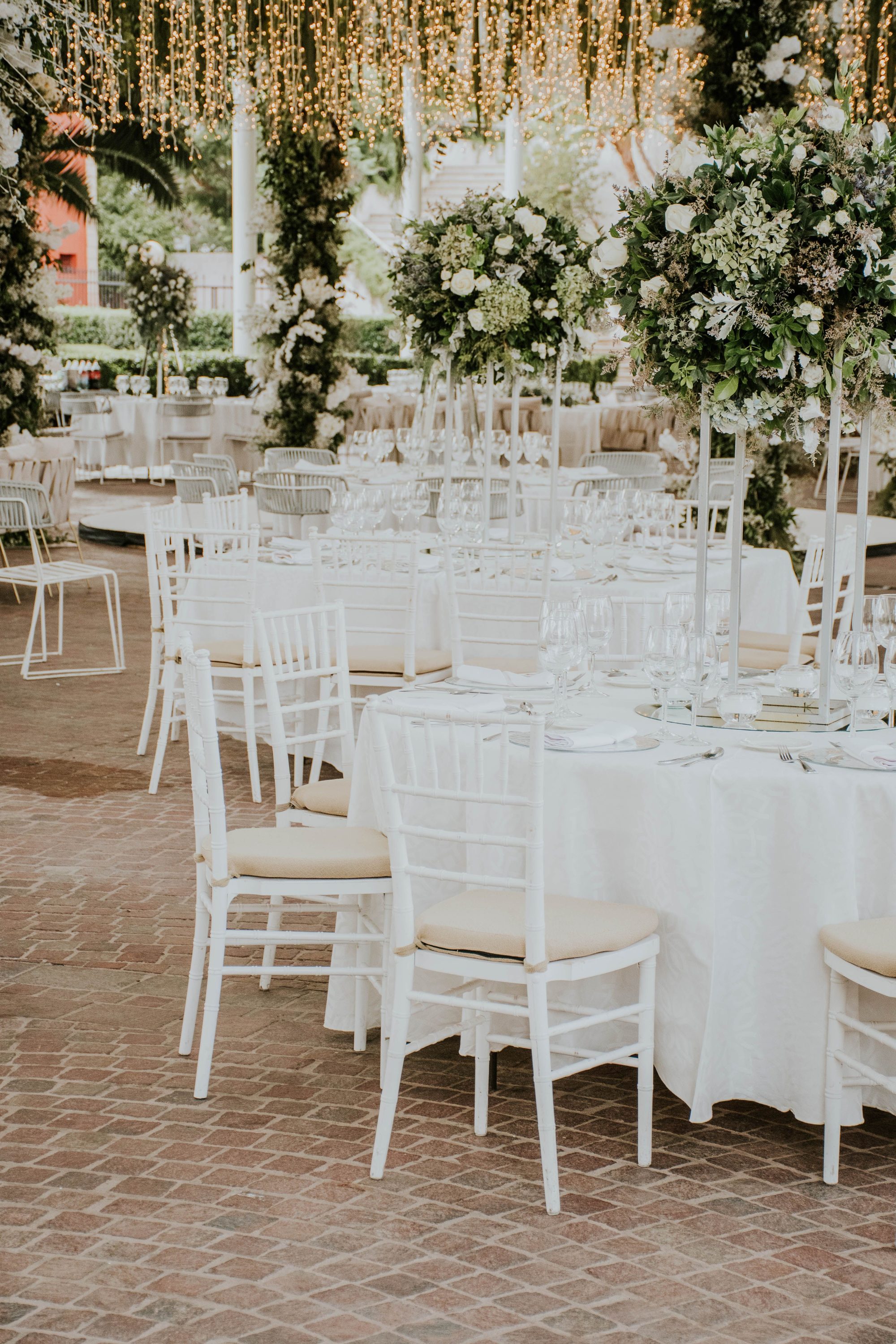 Outdoor wedding reception at golden hour with string lights overhead, tall white floral centerpieces, and white chiavari chairs